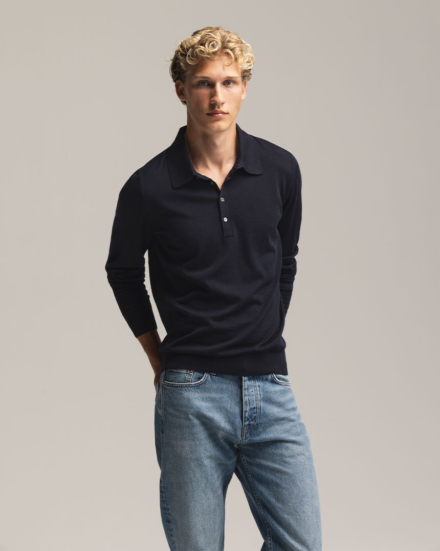 En person med lockigt hår bär en mörk tröja och jeans mot neutral bakgrund.