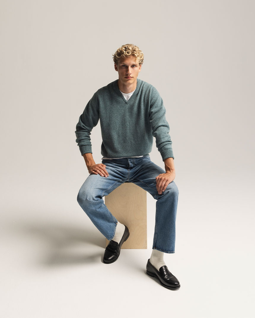 Person sitter på en trälåda, klädd i jeans, grön tröja, svarta loafers och vita strumpor, i en ljus miljö.