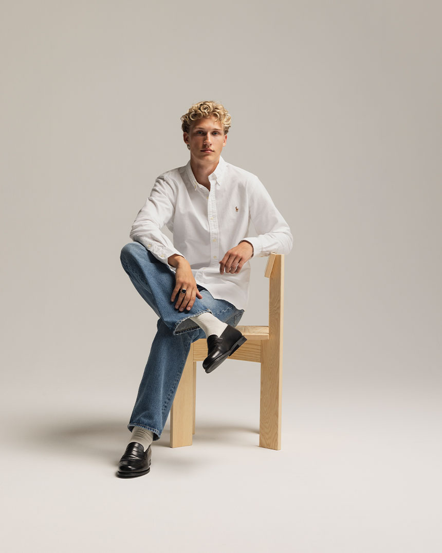 En person i vit skjorta och jeans sitter på en trästol mot en neutral bakgrund.