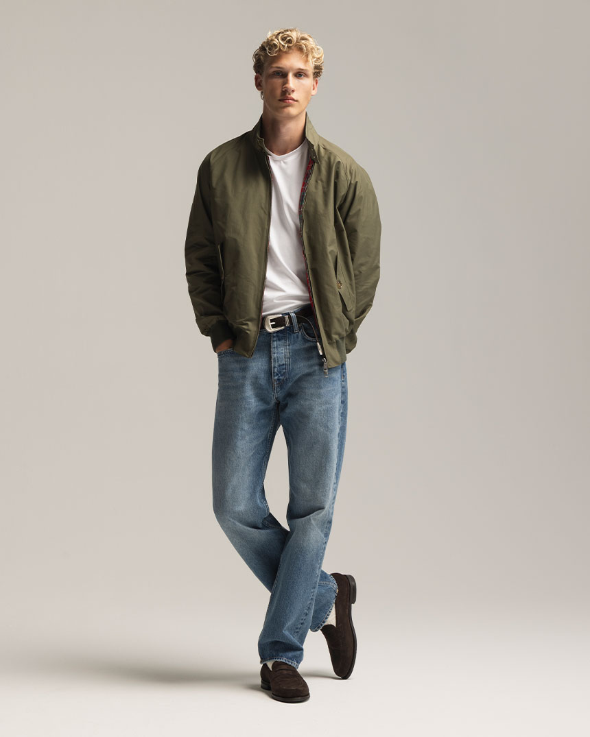 En person står i avslappnad pose, klädd i grön jacka, vit t-shirt, blå jeans och mörka skor.