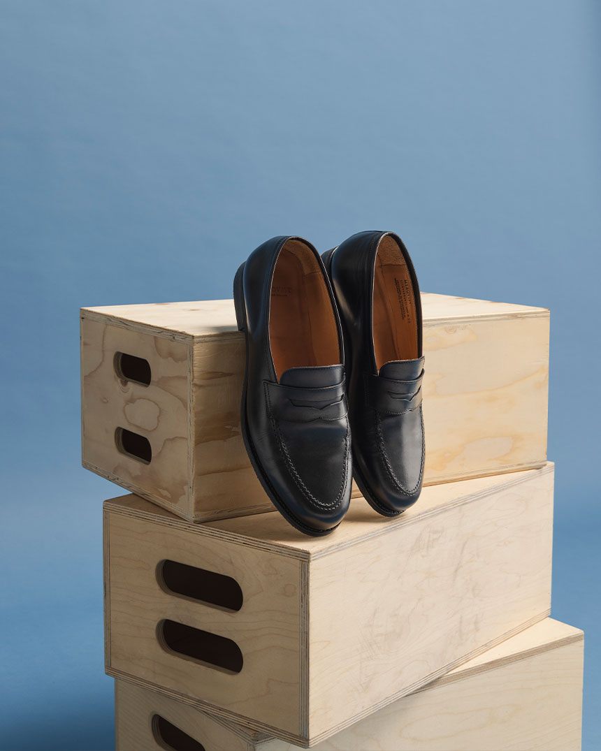 Ett par svarta loafers står på staplade träboxar mot en ljusblå bakgrund.